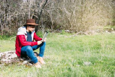 Girişimci serbest çalışan şapkalı bir adam, sahada dinlenirken tabletine bakıyor, bir taşın üzerinde oturuyor. İş kavramı, keyif, rahatlama, İnternet, teknoloji ve bağlantı.