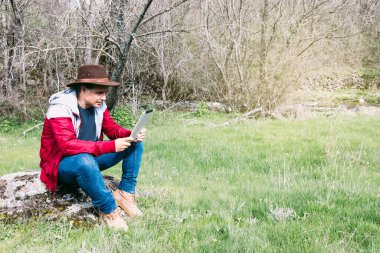 Girişimci serbest çalışan şapkalı bir adam, sahada dinlenirken tabletine bakıyor, bir taşın üzerinde oturuyor. İş kavramı, keyif, rahatlama, İnternet, teknoloji ve bağlantı.