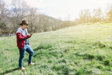 Girişimci, şapkalı, otlakta dinlenirken cep telefonuna bakan serbest meslek sahibi bir adam. İş kavramı, keyif, rahatlama, İnternet, teknoloji ve bağlantı.