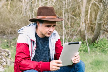 Girişimci serbest çalışan şapkalı bir adam, sahada dinlenirken tabletine bakıyor, bir taşın üzerinde oturuyor. İş kavramı, keyif, rahatlama, İnternet, teknoloji ve bağlantı.