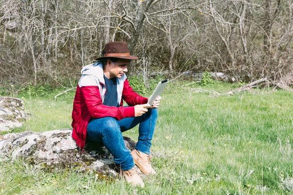 Girişimci serbest çalışan şapkalı bir adam, sahada dinlenirken tabletine bakıyor, bir taşın üzerinde oturuyor. İş kavramı, keyif, rahatlama, İnternet, teknoloji ve bağlantı.
