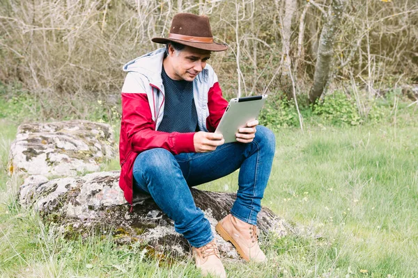 Girişimci serbest çalışan şapkalı bir adam, sahada dinlenirken tabletine bakıyor, bir taşın üzerinde oturuyor. İş kavramı, keyif, rahatlama, İnternet, teknoloji ve bağlantı.