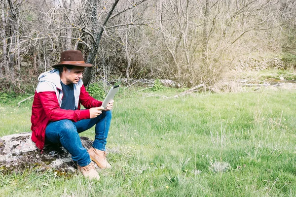 Girişimci serbest çalışan şapkalı bir adam, sahada dinlenirken tabletine bakıyor, bir taşın üzerinde oturuyor. İş kavramı, keyif, rahatlama, İnternet, teknoloji ve bağlantı.