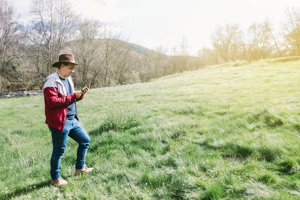 Girişimci, şapkalı, otlakta dinlenirken cep telefonuna bakan serbest meslek sahibi bir adam. İş kavramı, keyif, rahatlama, İnternet, teknoloji ve bağlantı.