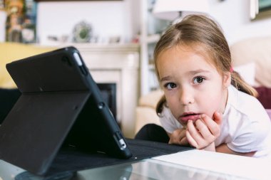 A little blonde girl in front of a tablet, looking at the camera in her living room. Concept of childhood, technology, learning, having fun, the Internet and connectivity.