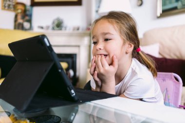A little blonde girl intently watching a video on a tablet in her living room. Concept of childhood, technology, learning, having fun, the Internet and connectivity.