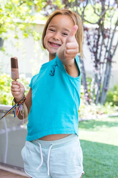 Unknown girl, wearing green t-shirt, holding a milk chocolate ice cream, on the street, in summer, raising her thumb. Ice cream, popsicle, eating, sweet and summer concept.