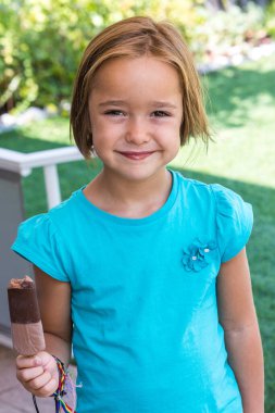 Unknown girl, wearing a green t-shirt, holding a milk chocolate ice cream, on the street, in summer, looking at the camera. Ice cream, popsicle, eating, sweet and summer concept.
