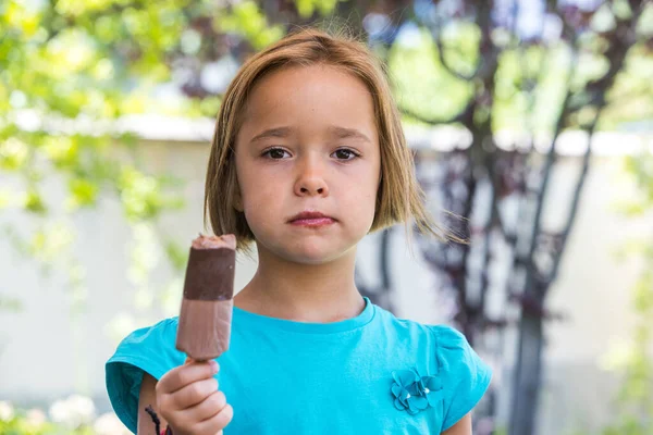 Unknown girl, wearing a green t-shirt, holding a milk chocolate ice cream, on the street, in summer, looking at the camera. Ice cream, popsicle, eating, sweet and summer concept.