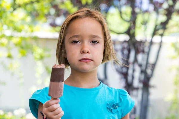 Unknown girl, wearing a green t-shirt, holding a milk chocolate ice cream, on the street, in summer, looking at the camera. Ice cream, popsicle, eating, sweet and summer concept.