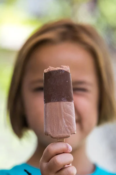 Unknown girl, wearing green shirt. out of focus, holding a milk chocolate ice cream, focused, on the street, in summer. Ice cream, popsicle, eating, sweet and summer concept.