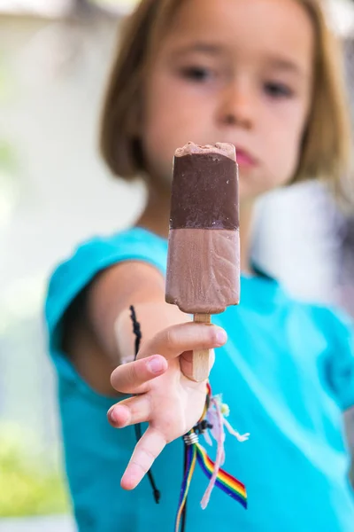 Unknown girl, wearing green shirt. out of focus, holding a milk chocolate ice cream, focused, on the street, in summer. Ice cream, popsicle, eating, sweet and summer concept.