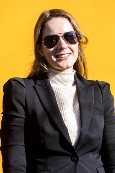 Executive entrepreneur woman, wearing black jacket and wool sweater and sunglasses, on yellow background in the street, with copy space, looking at camera and smiling. Entrepreneur woman concept.