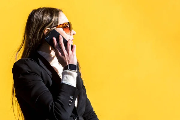 Executive entrepreneur woman, wearing black jacket and wool sweater and sunglasses, on yellow background in the street, with copy space, talking on the mobile. Entrepreneur woman concept.