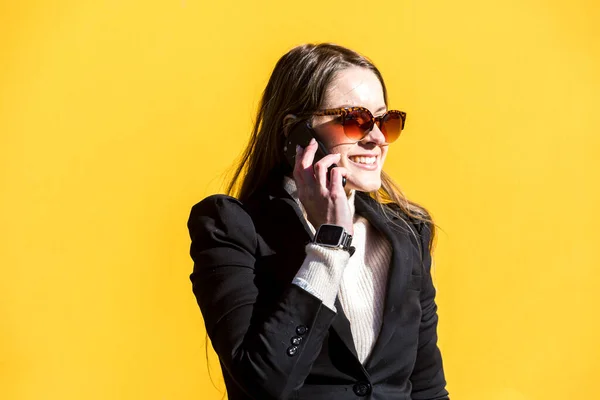 Executive entrepreneur woman, wearing black jacket and wool sweater and sunglasses, on yellow background in the street, with copy space, talking on the mobile. Entrepreneur woman concept.