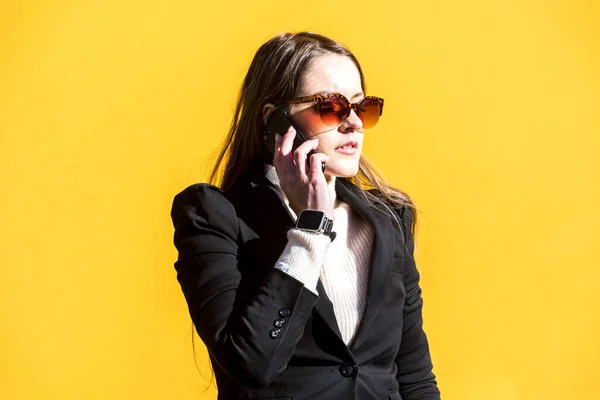 Executive entrepreneur woman, wearing black jacket and wool sweater and sunglasses, on yellow background in the street, with copy space, talking on the mobile. Entrepreneur woman concept.