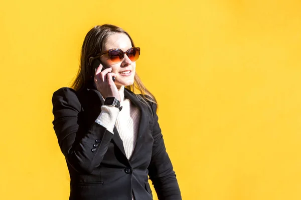 Executive entrepreneur woman, wearing black jacket and wool sweater and sunglasses, on yellow background in the street, with copy space, talking on the mobile. Entrepreneur woman concept.