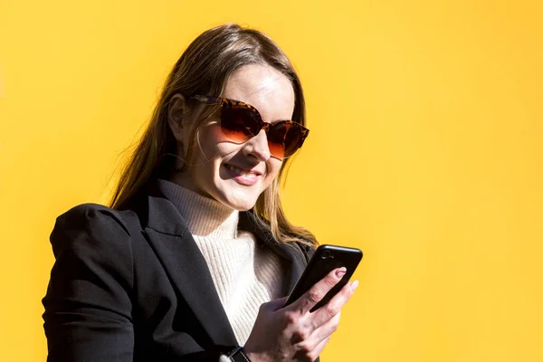 Executive entrepreneur woman, wearing black jacket and wool sweater and sunglasses, on yellow background in the street, with copy space, looking at mobile. Entrepreneur woman concept.