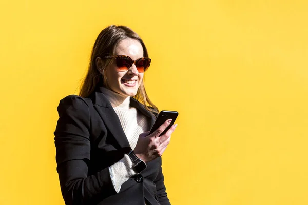 Executive entrepreneur woman, wearing black jacket and wool sweater and sunglasses, on yellow background in the street, with copy space, looking at mobile. Entrepreneur woman concept.