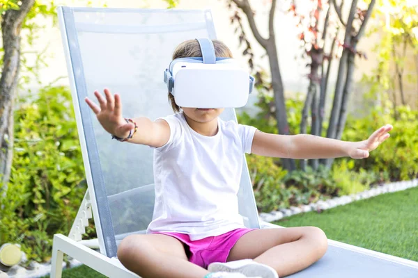 Little girl with virtual reality glasses, trying to touch something in a virtual way, sitting on a sun lounger in the garden of her house. Metaverse, VR, game, digital and simulation concept.