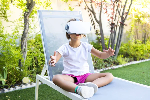 Little girl with virtual reality glasses, trying to touch something in a virtual way, sitting on a sun lounger in the garden of her house. Metaverse, VR, game, digital and simulation concept.