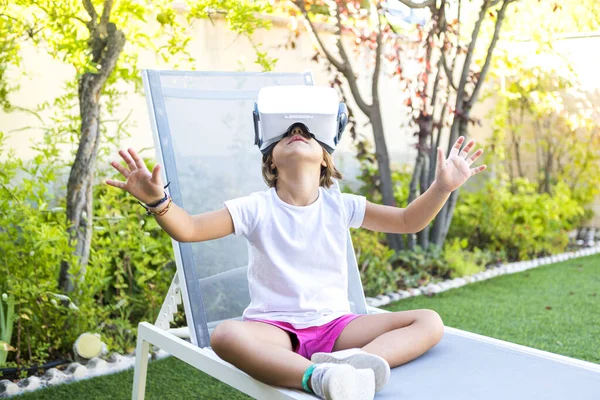 Little girl wearing virtual reality goggles, looking up, trying to touch something in a virtual way, sitting on a deckchair in the garden of her house. Metaverse, VR, game, simulation concept.