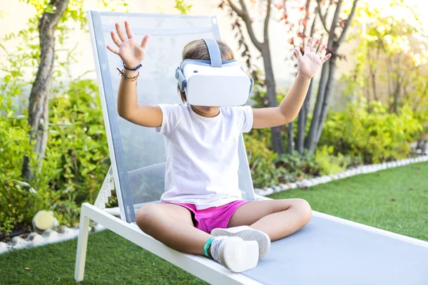 Little girl with virtual reality glasses, trying to touch something in a virtual way, sitting on a sun lounger in the garden of her house. Metaverse, VR, game, digital and simulation concept.