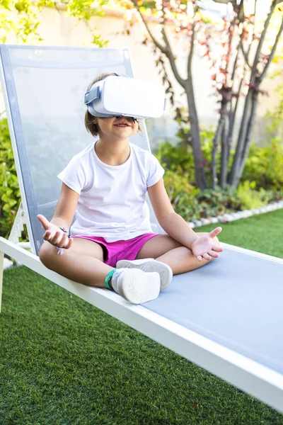 Little girl with virtual reality glasses, trying to touch something in a virtual way, sitting on a sun lounger in the garden of her house. Metaverse, VR, game, digital and simulation concept.