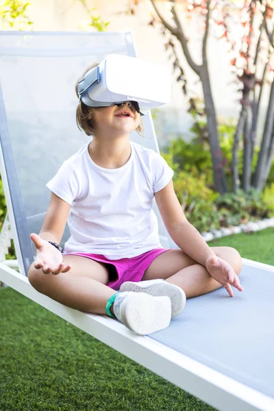 Little girl with virtual reality glasses, trying to touch something in a virtual way, sitting on a sun lounger in the garden of her house. Metaverse, VR, game, digital and simulation concept.