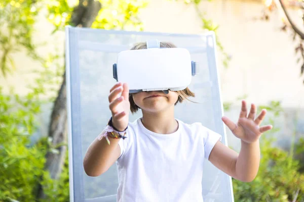 Little girl with virtual reality glasses, trying to touch something in a virtual way, sitting on a sun lounger in the garden of her house. Metaverse, VR, game, digital and simulation concept.