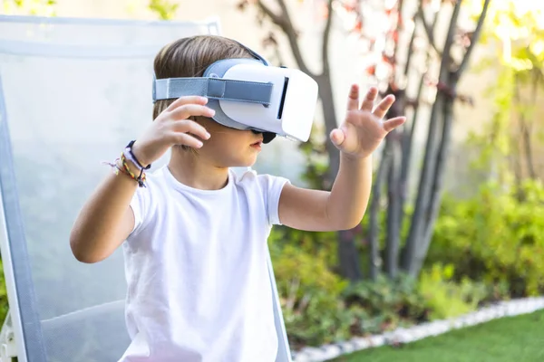 Little girl with virtual reality glasses, looking to the right, trying to touch something in a virtual way, sitting on a sun lounger in the garden of her house. Metaverse, VR, game, digital concept.