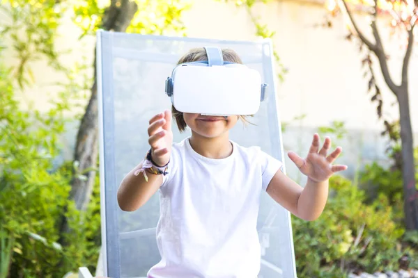 Little girl with virtual reality glasses, trying to touch something in a virtual way, sitting on a sun lounger in the garden of her house. Metaverse, VR, game, digital and simulation concept.