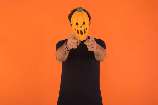 Person with pumpkin mask celebrating Halloween, pointing at camera, on orange background. Concept of celebration, All Souls' Day and All Saints' Day.