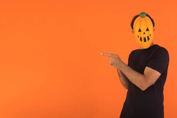 Person with pumpkin mask celebrating Halloween, pointing to the side with fingers, on orange background. Concept of celebration, All Souls\' Day and All Saints\' Day.