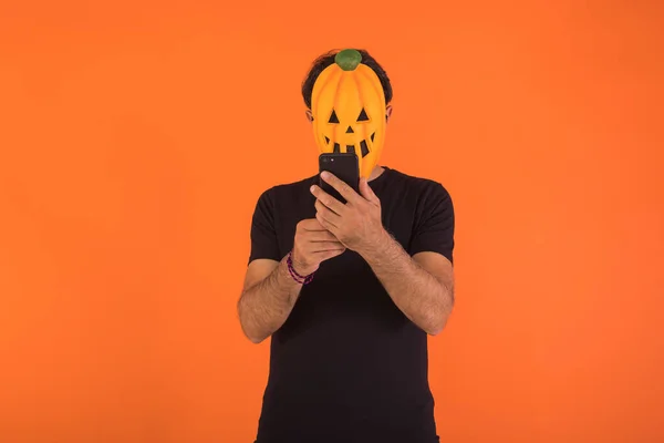 Person with pumpkin mask celebrating Halloween, looking at his mobile phone, on orange background. Concept of celebration, All Souls\' Day and All Saints\' Day.