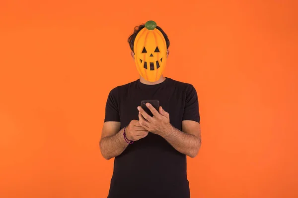 Person with pumpkin mask celebrating Halloween, looking at his mobile phone, on orange background. Concept of celebration, All Souls\' Day and All Saints\' Day.