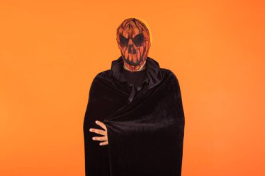 Balkabağı balkabağı maskesi ve siyah kapüşonlu pelerin takan, kollarını kavuşturmuş, Cadılar Bayramı 'nı turuncu arka planda kutlayan biri. Kutlama konsepti, Ruhlar Günü ve Azizler Günü..