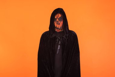 Balkabağı balkabağı maskesi ve siyah kapüşonlu pelerin takan kişi Cadılar Bayramı 'nı turuncu arka planda kutluyor. Kutlama konsepti, Ruhlar Günü ve Azizler Günü..