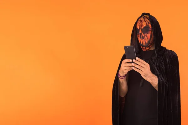 Person wearing Jack o Lantern pumpkin mask and black hooded cape, consulting his mobile phone, celebrating Halloween, on orange background. Celebration concept, All Souls\' Day and All Saints\' Day.