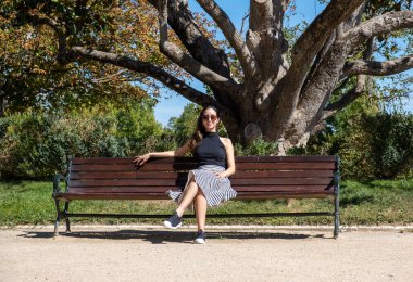 Güneş gözlüklü, yaz kıyafetleriyle genç, şık bir kadın parkta bir bankta oturmuş dinleniyor.