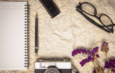 Gişede film kamerası, kalem, film rulosu ve mor çiçeklerle dolu boş bir sayfası olan defter ofisi. Üst görünüm buruşmuş kağıt vintage ton.