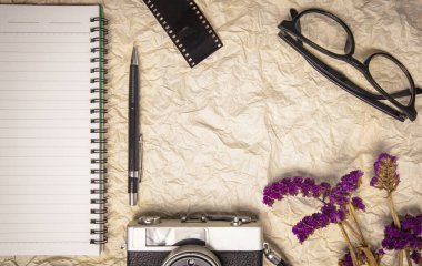 Gişede film kamerası, kalem, film rulosu ve mor çiçeklerle dolu boş bir sayfası olan defter ofisi. Üst görünüm buruşmuş kağıt vintage ton.