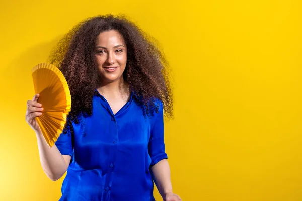 Afro girl smiling using a fan. Copy space on white background. Heat concept on yellow background