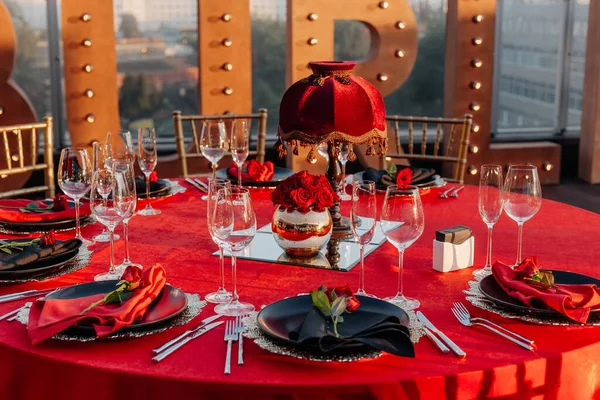 Guests table setting for banquet in black, red and gold style. Elegant dinner: decor, tablecloth, plates with napkins and fresh roses, glasses, cutlery. Themed party celebration on the roof, outdoor