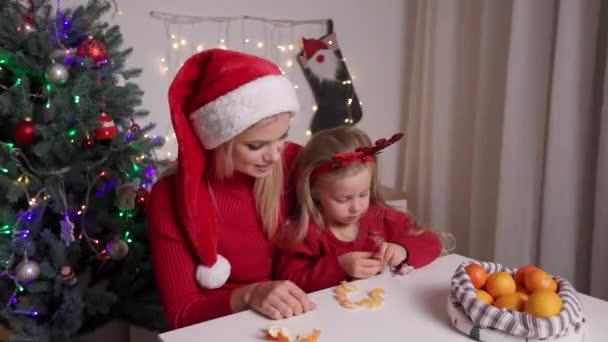 妈妈戴着红色桑塔帽子，在圣诞树下吃橙子的滑稽孩子. — 图库视频影像