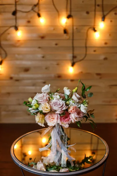 Λεπτή και πολύ όμορφη γαμήλια ανθοδέσμη από τριαντάφυλλα, πράσινο, ευστόμα στέκεται στο τραπέζι καθρέφτη. Νυφικά μοντέρνα λουλούδια σε παστέλ χρώματα στο φόντο των λαμπτήρων φωτισμού βράδυ. — Φωτογραφία Αρχείου