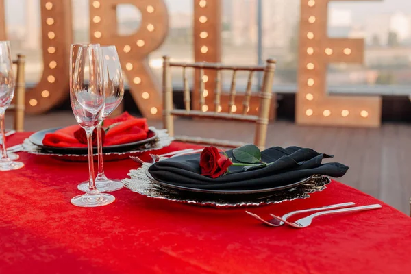 Guests table setting for banquet in black, red and gold style. Elegant dinner: decor, tablecloth, plates with napkins and fresh roses, glasses, cutlery. Themed party celebration on the roof, outdoor.