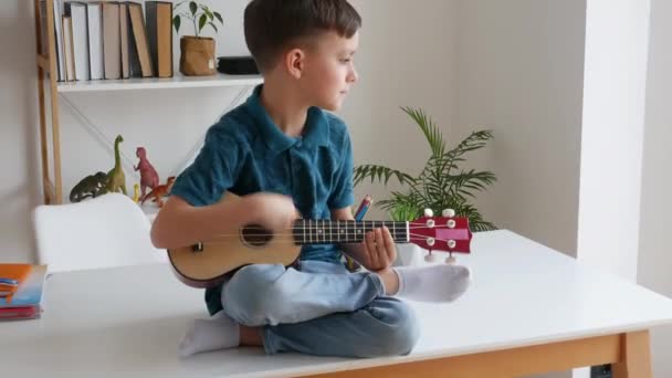 Niño talentoso jugando ukelele soprano sentado en el escritorio. Niño preescolar aprendiendo guitarra en el ocio. Concepto de educación infantil y pasatiempo musical. — Vídeos de Stock