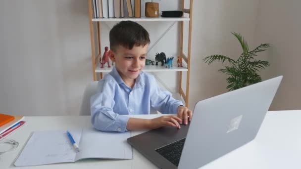 Ребенок через ноутбук разговаривает с учителем во время онлайн обучения дома. Электронное обучение в начальной школе через видеосвязь. Умный мальчик делает домашнее задание с помощью интернета и гаджета. — стоковое видео