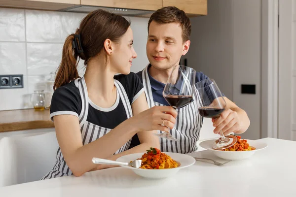 Valentine day romantic dinner at home. Happy couple smiling, clinking glasses and eating pasta with wine at kitchen. Concept of domestic lifestyle, love, happy marriage and togetherness — Stock Photo, Image